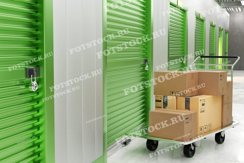 На фотографии изображена тележка с картонными коробками, расположенная в коридоре складского помещения с зелеными дверями. Видны запертые боксы, что символизирует безопасное хранение личных вещей в арендованных складских ячейках.